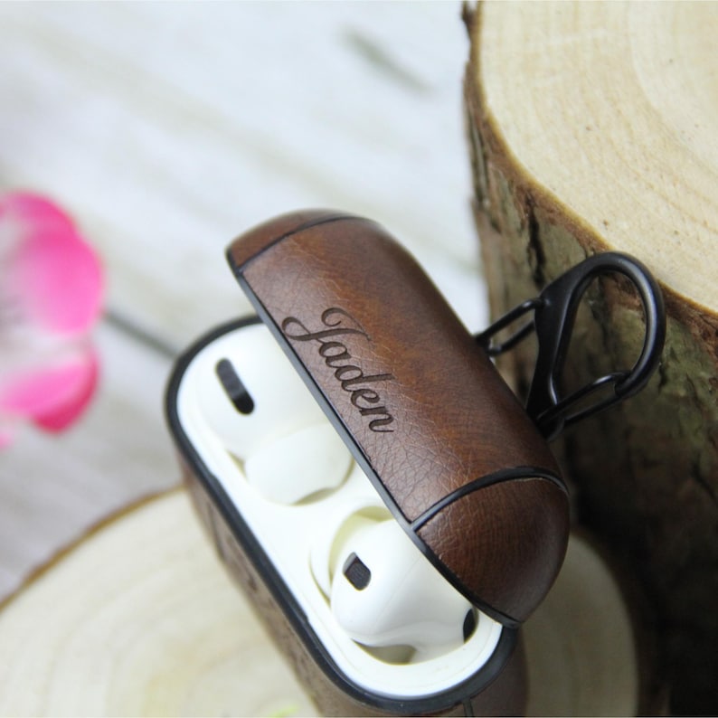 May include: Brown leather AirPod case with the name "Jaden" engraved on the lid. The case is open, revealing white earbuds inside. A black carabiner clip is attached to the side. The case is on a wooden surface.