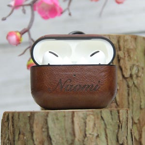 May include: A brown leather protective case for wireless earbuds with the name Naomi engraved on the front. The case is open, revealing white earbuds inside. The exterior has a textured finish, and the interior lining is white plastic.