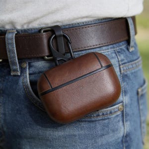 Peut inclure: Un &eacute;tui en cuir marron pour AirPods Pro avec une bordure noire et un mousqueton noir, attach&eacute; &agrave; une ceinture marron. L'&eacute;tui est dans la poche d'un jean bleu.