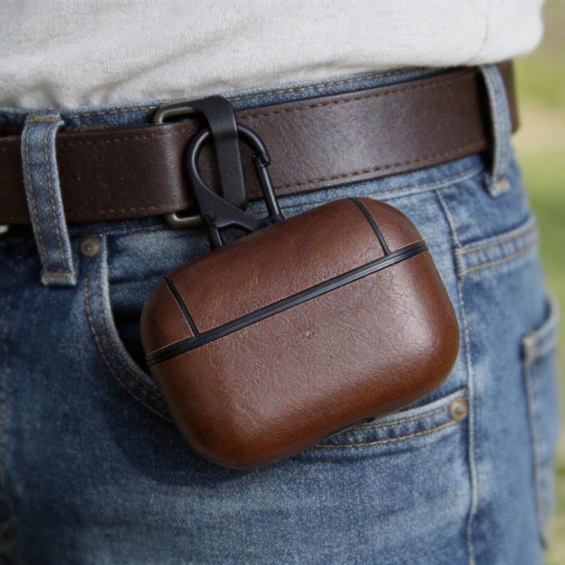 Peut inclure: Un &eacute;tui en cuir marron pour AirPods Pro avec une bordure noire et un mousqueton noir, attach&eacute; &agrave; une ceinture marron. L'&eacute;tui est dans la poche d'un jean bleu.