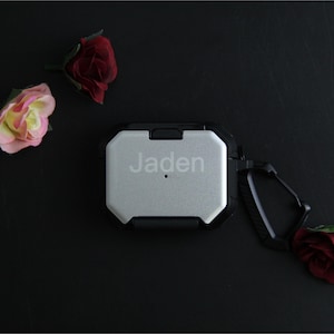 Puede incluir: Funda protectora plateada y negra para auriculares inal&aacute;mbricos, con el nombre "Jaden" grabado en la parte delantera. La funda tiene un mosquet&oacute;n y est&aacute; rodeada de rosas artificiales sobre una superficie negra.