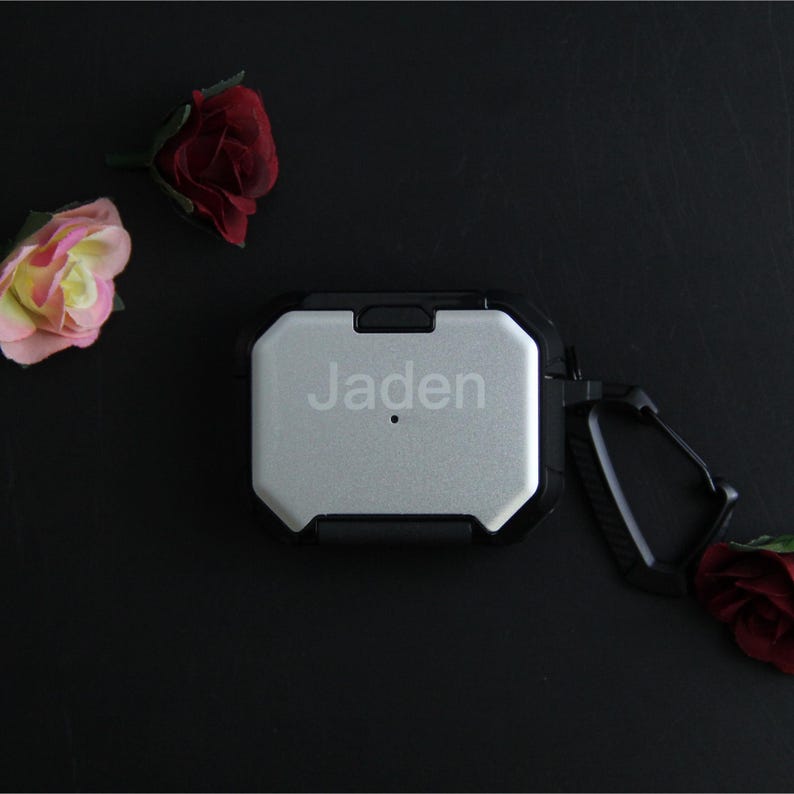 Puede incluir: Funda protectora plateada y negra para auriculares inal&aacute;mbricos, con el nombre "Jaden" grabado en la parte delantera. La funda tiene un mosquet&oacute;n y est&aacute; rodeada de rosas artificiales sobre una superficie negra.