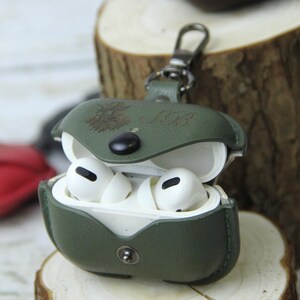Op de afbeelding: Een groene leren AirPods Pro-hoes met een zilveren sluiting, open om witte oortelefoons te onthullen. De hoes heeft een knoopsluiting en is voorzien van initialen en een ontwerp. De hoes ligt op een houten oppervlak.