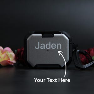 Puede incluir: Una funda protectora gris y negra para auriculares inal&aacute;mbricos. La funda tiene el nombre "Jaden" grabado en la parte delantera, con una flecha que apunta al texto "Your Text Here". La funda tiene un mosquet&oacute;n negro.