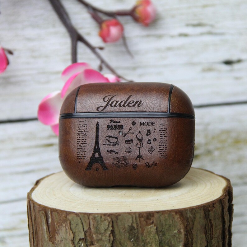 May include: Brown leather AirPods case with the name "Jaden" engraved on top. The case features a design with the Eiffel Tower, coffee cup, scissors, and the words "Paris" and "Mode". The case is sitting on a wooden surface.