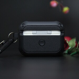 May include: A black earbud case with a textured finish, a circular button, and a metal loop for a keyring. The case is designed to protect wireless earbuds. The case is a dark, matte black.
