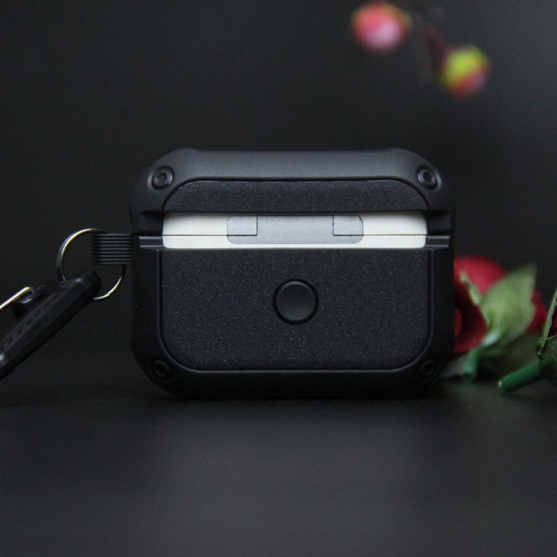 May include: A black earbud case with a textured finish, a circular button, and a metal loop for a keyring. The case is designed to protect wireless earbuds. The case is a dark, matte black.