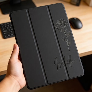 May include: Black tablet case with a rose design and the name "Isabella" engraved on the front. The case is folded and has a magnetic closure. The rose is detailed with leaves and petals.
