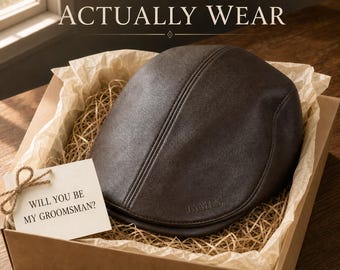 May include: A dark brown flat cap with the name JAMES embossed on the side, presented in a cardboard gift box with paper filler and a small white card tied with twine that reads WILL YOU BE MY GROOMSMAN? The text A GIFT HE'LL ACTUALLY WEAR is visible.