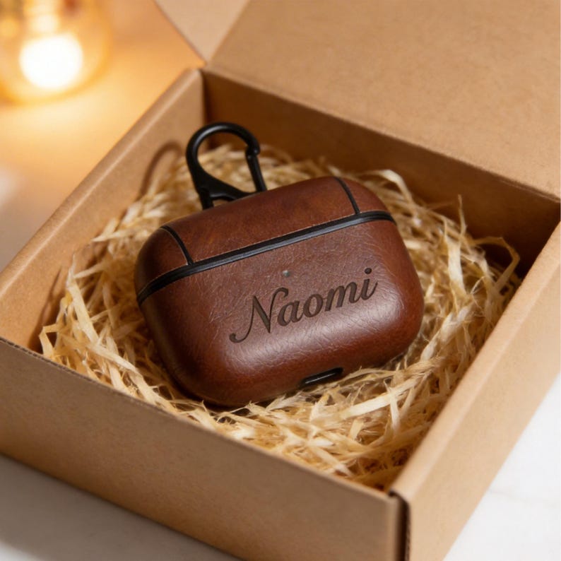 Peut inclure: Un &eacute;tui AirPod en cuir marron avec le nom "Naomi" grav&eacute; sur le devant. L'&eacute;tui a une bordure noire et un mousqueton noir. Il est pos&eacute; dans une bo&icirc;te en carton remplie de paille.