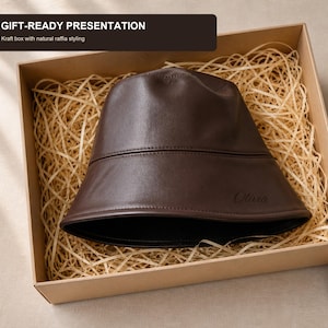 Op de afbeelding: Een donkerbruine bucket hat met een gladde afwerking en de naam Olivia in reliëf aan de zijkant. De hoed wordt gepresenteerd in een rechthoekige kraftdoos gevuld met natuurlijk raffia, met de tekst GIFT-READY PRESENTATION bovenaan.
