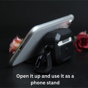 Puede incluir: Una funda protectora negra y gris para auriculares inal&aacute;mbricos con un mosquet&oacute;n, que sostiene un smartphone plateado. La imagen incluye el texto "Open it up and use it as a phone stand."