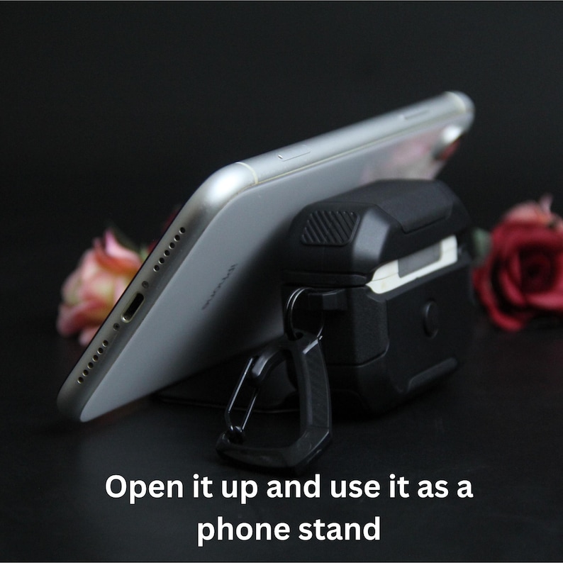 Puede incluir: Una funda protectora negra y gris para auriculares inal&aacute;mbricos con un mosquet&oacute;n, que sostiene un smartphone plateado. La imagen incluye el texto "Open it up and use it as a phone stand."