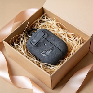 Puede incluir: Una funda para AirPods gris oscuro con un mosquetón negro, presentada en una caja de cartón llena de paja. La funda tiene grabado el nombre "Naomi", junto con un gráfico de dos figuras. Una cinta rosa está drapeada alrededor de la caja.