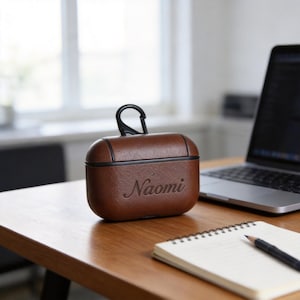 Peut inclure: Un &eacute;tui en cuir marron pour AirPods avec le nom "Naomi" en relief sur le devant. L'&eacute;tui a une bordure noire et un mousqueton noir. L'&eacute;tui est pos&eacute; sur un bureau en bois &agrave; c&ocirc;t&eacute; d'un ordinateur portable et d'un bloc-notes.