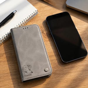 Peut inclure: Un étui de téléphone portefeuille gris avec une illustration gravée au laser d'une tête de chien et le nom Naomi imprimé en dessous. L'étui présente un petit accent métallique circulaire dans le coin inférieur droit et une finition mate.