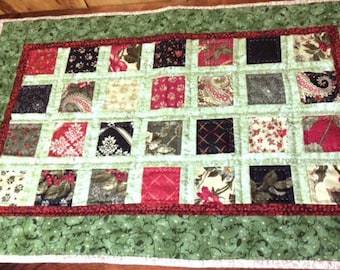 May include: A green and red quilted table runner with a patchwork design. The runner is made up of small squares of fabric in various patterns and colors. The runner has a white border and a red and green border.
