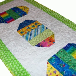May include: A white table runner with a green border and three colorful patchwork egg designs. The eggs are made of different fabrics with stripes, polka dots, and floral patterns.