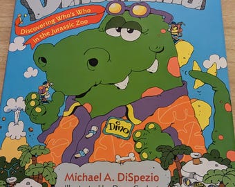 May include: A green dinosaur with orange, purple, and white clothing, wearing glasses and a name tag that says "Dino". The book cover features the text "Dinomania: Discovering Who's Who in the Jurassic Zoo" and the author's name, "Michael A. DiSpezio".
