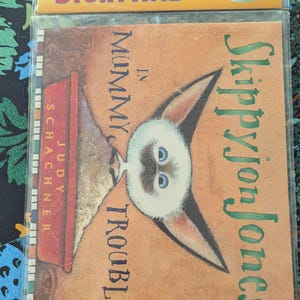 Puede incluir: Un libro y CD de Puffin Storytime titulado "Skippyjon Jones in Mummy Trouble" de Judy Schachner. La portada presenta un gato de dibujos animados con orejas grandes y ojos azules sobre un fondo naranja.