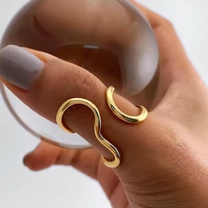 May include: A gold-colored ring with a unique, flowing design. The ring wraps around two fingers, with one part of the ring curving in a wave-like shape. The other part of the ring is a simple curved band. The ring is a modern piece of jewelry.