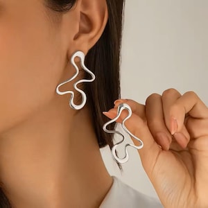 May include: Silver-toned abstract earrings. One earring is worn on the ear, while the other is held in the hand. The earrings have a fluid, irregular shape, with a smooth, reflective surface. The design is modern and minimalist.