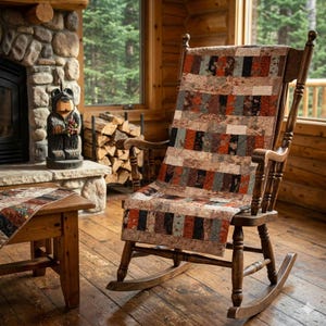 Puede incluir: Una mecedora de madera con una colcha de retazos sobre ella. La colcha presenta un patrón rectangular en tonos marrones, rojos y negros. La silla está en un entorno de cabaña rústica con chimenea y mesa de madera.