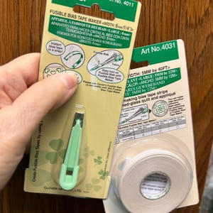 May include: Two Clover brand sewing tools are displayed. One package contains a green and silver bias tape maker, and the other holds a roll of fusible bias tape. The packaging includes text in multiple languages, including "FUSIBLE BIAS TAPE MAKER" and "WIDTH: 6mm (1/4")".