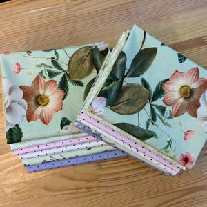 May include: Two stacks of floral fabric with a light green background. The fabric features large, peach-colored flowers, white flowers, and green leaves. Several smaller patterned fabrics in pink, white, and purple are stacked below.