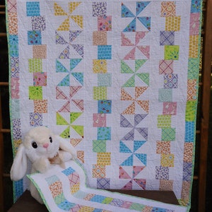 May include: A patchwork quilt featuring a pinwheel pattern with various pastel-colored fabric squares in yellow, pink, blue, and green, bordered by a white frame. A plush white rabbit toy is positioned in front of the quilt on a wooden surface.