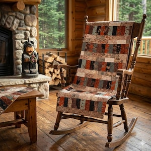 May include: A wooden rocking chair with a patchwork quilt draped over it. The quilt features rectangular blocks in shades of brown, red, and black. The chair is in a rustic cabin setting with a fireplace and wooden table.