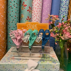 May include: A display of colorful embroidery scissors in pink, teal, and blue, with a vintage sign that reads "Vintage Stork Scissors." The scissors are on a clear acrylic stand, with rolls of patterned fabric in the background.