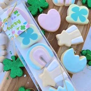 May include: A clear package of decorated sugar cookies with a St. Patrick's Day theme. The cookies are shaped like rainbows, shamrocks, and stars, and are decorated with pastel colors. The package has a label that says "Lucky Charms".