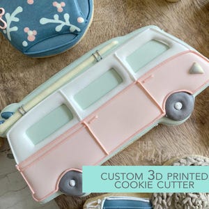 May include: A pink, white, and light blue camper van cookie cutter with a surfboard on top. The cookie cutter has a rectangular shape with rounded edges and a gray wheel. The text "CUSTOM 3D PRINTED COOKIE CUTTER" is visible.