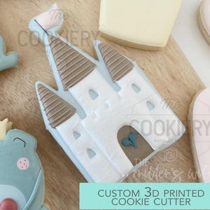 May include: A white castle-shaped cookie cutter with brown roof details and a blue heart on the door. The text "CUSTOM 3D PRINTED COOKIE CUTTER" is visible. Other cookies are visible in the background.