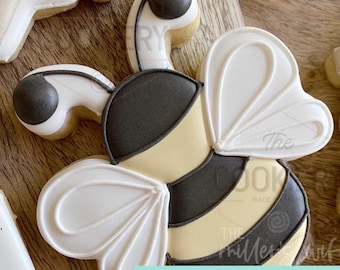 May include: A bee-shaped cookie cutter with black, white, and yellow icing. The cookie cutter has a detailed design with the text "CUSTOM 3D PRINTED COOKIE CUTTER" in a teal rectangle.