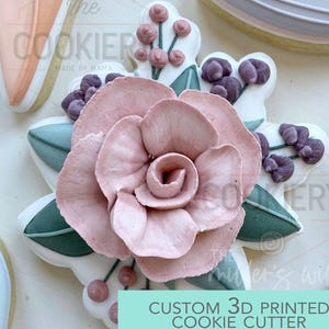 May include: A custom 3D printed cookie cutter in the shape of a pink rose with green leaves and purple berries. The cookie cutter is surrounded by other decorated cookies. Text on the image reads "CUSTOM 3D PRINTED COOKIE CUTTER".