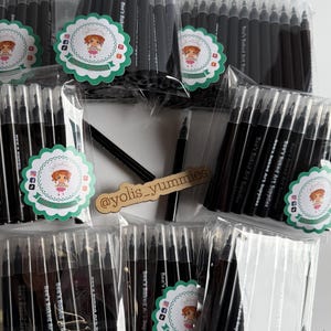 May include: Multiple packages of black art pens are displayed, each sealed in clear plastic. The pens are labeled with the text "Bee's Baked Art Supplies" and feature a logo. A wooden sign with the text "@yolis_yummies" is also visible.