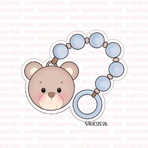 May include: A cookie cutter in the shape of a baby's pacifier with a teddy bear face. The pacifier is light blue with a round ring and a beaded design. The teddy bear has a light brown face with a pink blush.