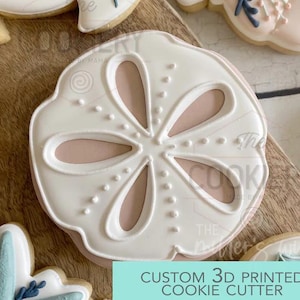 May include: A white, sand dollar-shaped cookie cutter with a light pink interior. The cookie cutter has a detailed design with small dots and lines. The text "CUSTOM 3D PRINTED COOKIE CUTTER" is in a light blue rectangle.