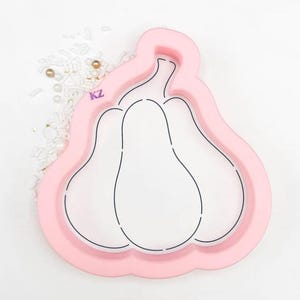 May include: A pink plastic cookie cutter in the shape of three pears. The cutter has a raised border and black outline details. The letters "KZ" are visible on the side. Sprinkles and gold beads are scattered around the cutter.
