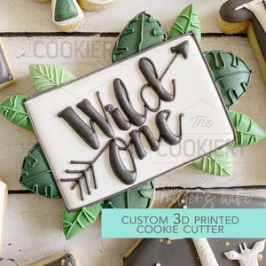 May include: A rectangular cookie with the words "Wild One" in dark gray script, with an arrow design. The cookie is surrounded by green leaf-shaped cookies. The text "CUSTOM 3D PRINTED COOKIE CUTTER" is at the bottom.