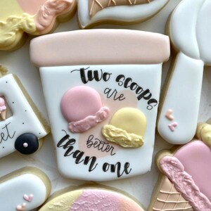 May include: A decorated sugar cookie shaped like an ice cream cup with the text "Two scoops are better than one." The cookie features two scoops of ice cream, one pink and one yellow, with a pink rim.