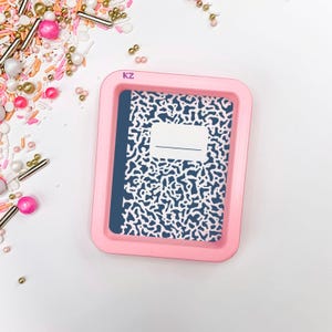 May include: A pink rectangular tray with a notebook design. The notebook has a blue cover and a white and blue pattern. The tray is surrounded by sprinkles and decorative beads.