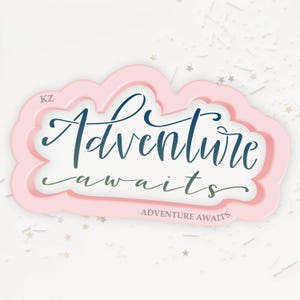 May include: A pink cloud-shaped cookie cutter with the words "Adventure awaits" in a cursive font. The text is in a gradient of blue and green. The words "Adventure awaits" are also printed in a smaller font at the bottom.