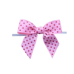 Op de afbeelding: Een roze strik met rode stippen. De strik is gemaakt van lint en heeft een klassieke vorm. Hij is bevestigd aan een transparante plastic stok, waarschijnlijk voor presentatie of bevestiging.