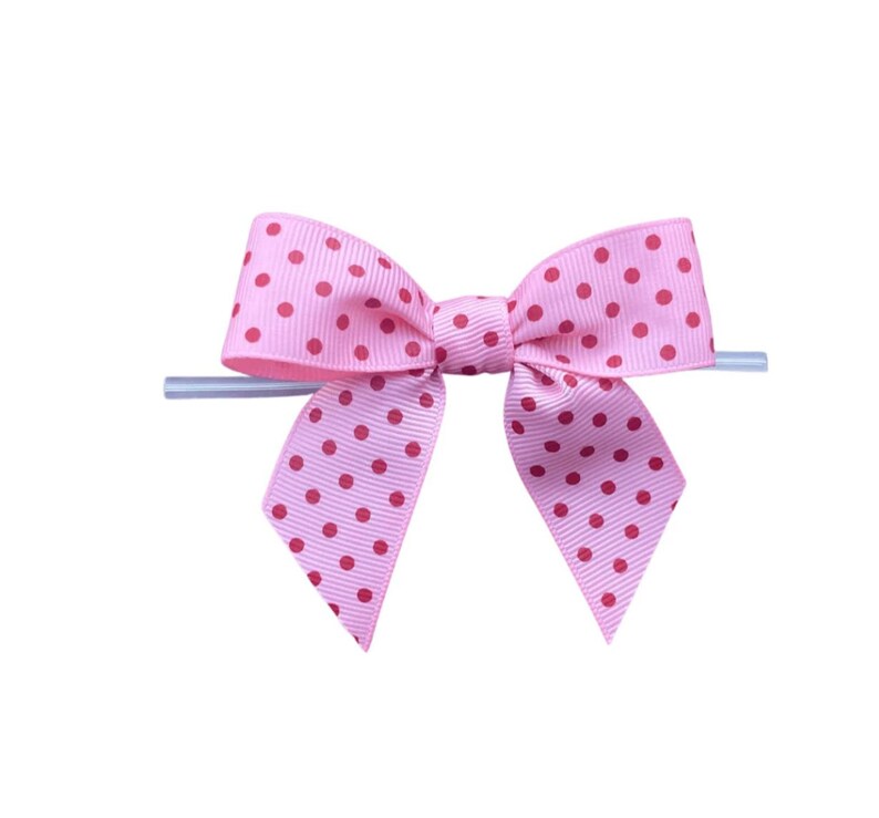 Op de afbeelding: Een roze strik met rode stippen. De strik is gemaakt van lint en heeft een klassieke vorm. Hij is bevestigd aan een transparante plastic stok, waarschijnlijk voor presentatie of bevestiging.