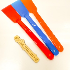 May include: Three colorful spatulas in red, blue, and orange are arranged diagonally. Each spatula has a rectangular head and a long handle with a hole. A wooden tag with the text "@yolis_yummies" is also present.