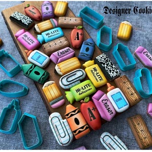 May include: A collection of school-themed cookies, including crayons, pencils, erasers, and apples, arranged on a wooden board. The cookies are decorated with vibrant colors and the text "HI-LITE delicious", "Glue", and "Eraser". Blue cookie cutters are also present.
