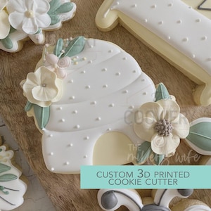 May include: White cookie cutter in the shape of a beehive, decorated with white dots and floral accents. The text "CUSTOM 3D PRINTED COOKIE CUTTER" is printed on a teal rectangle. Other decorated cookies are visible.